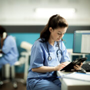 Nurse in blue scrubs reviewing notes on a tablet in a hospital.